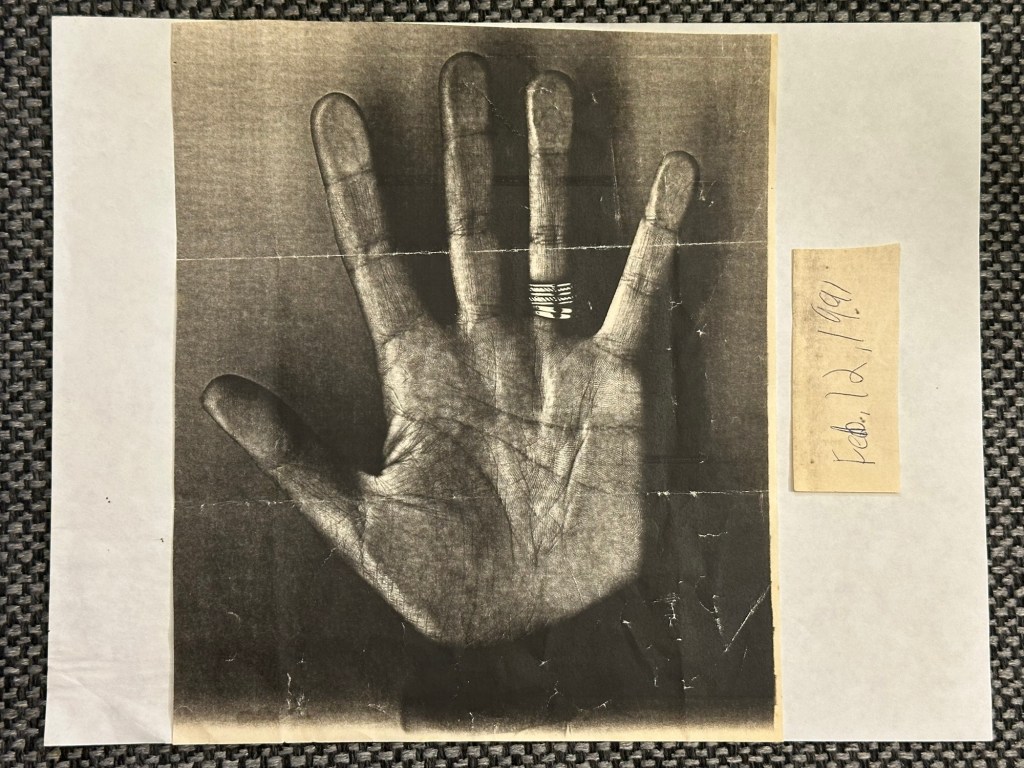 A black and white xerox image of a woman's open palm wearing a wedding band and an engagement ring with a piece of paper next to it saying Feb. 12, 1991.