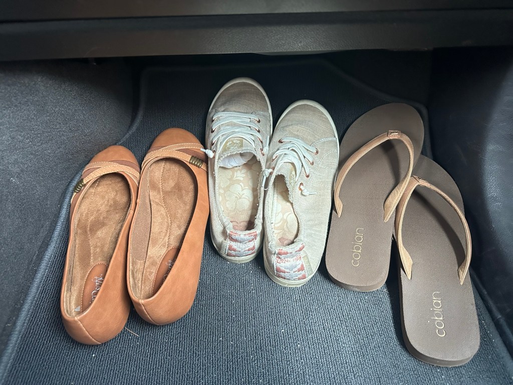 Three pairs of footwear on the floor of a car: tan flats, beige sneakers, and tan flip-flops.